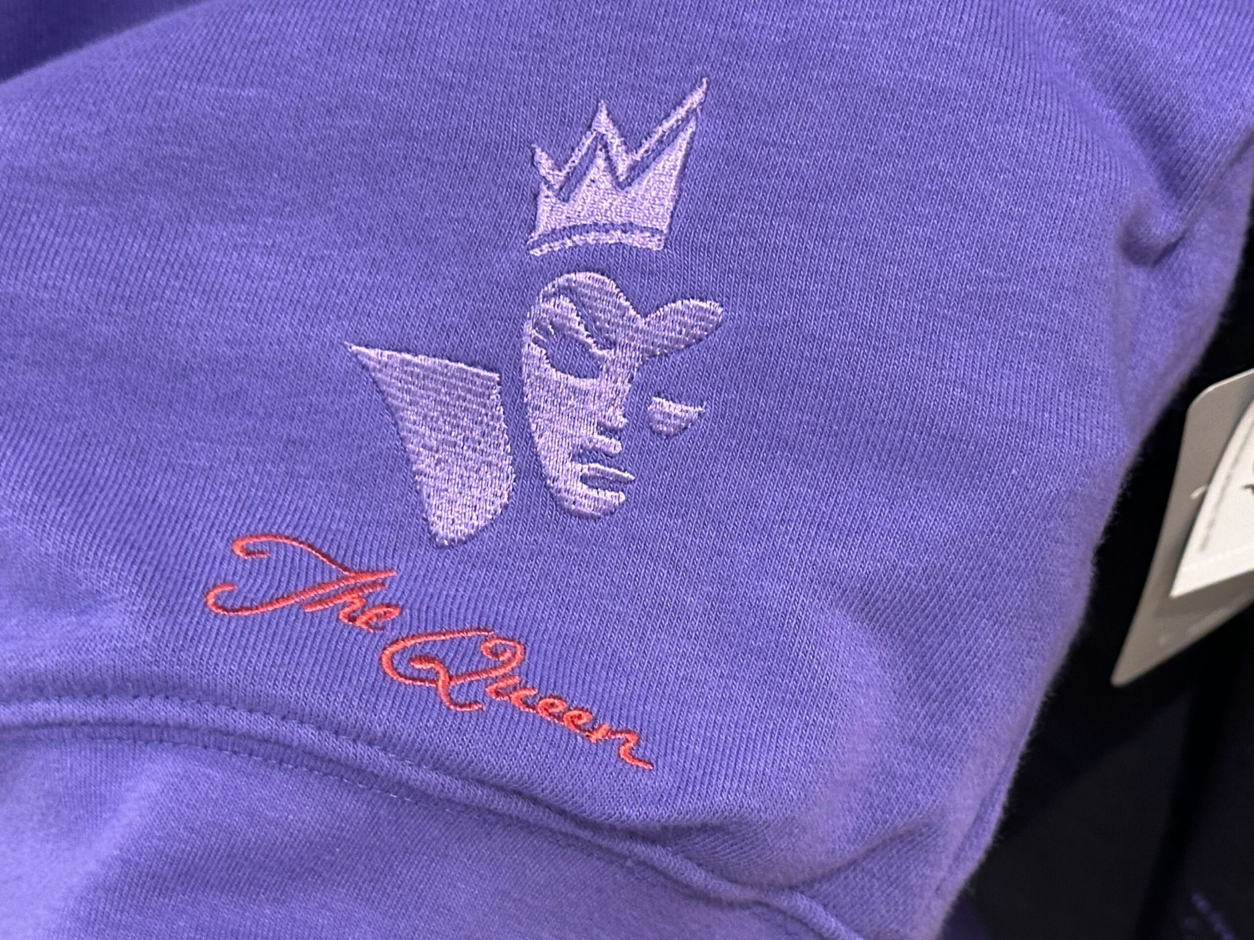 Tissu violet avec visage abstrait brodé portant une couronne rouge et la mention 'The Queen'
